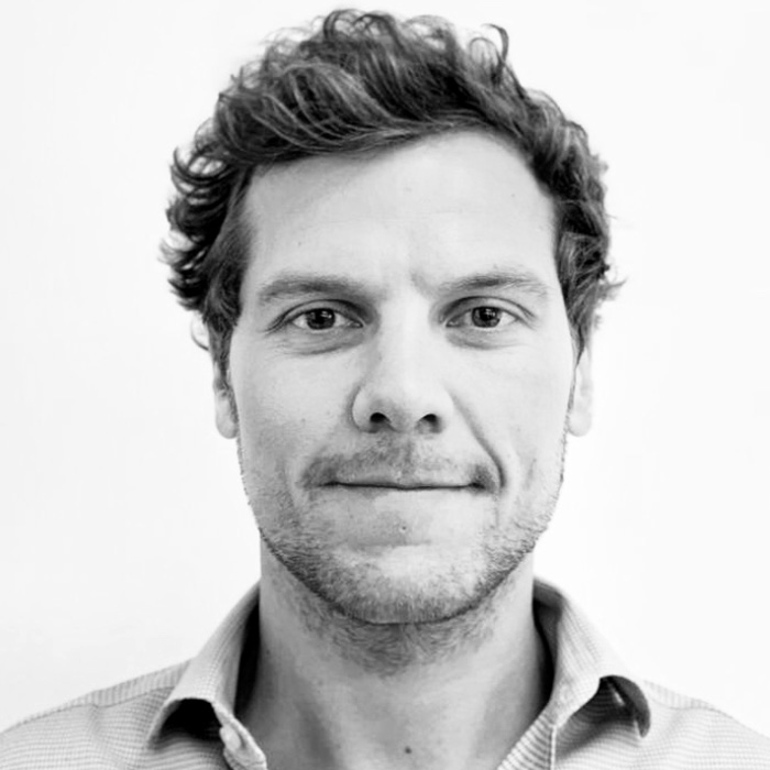 Close-up black and white portrait of a man with short curly hair, light stubble, and a collared shirt, looking directly at the camera with a neutral expression.