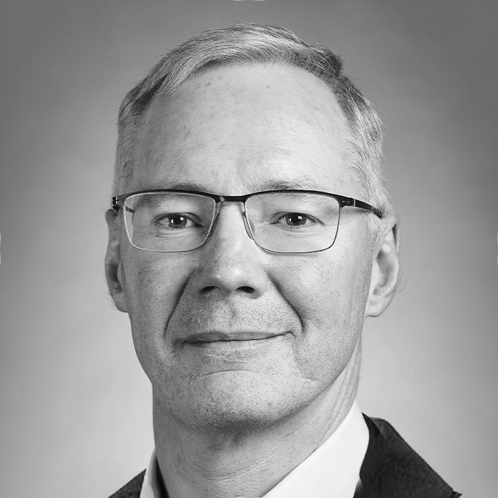Black and white headshot of a middle-aged man wearing glasses, a suit jacket, and a collared shirt, smiling slightly against a plain background.